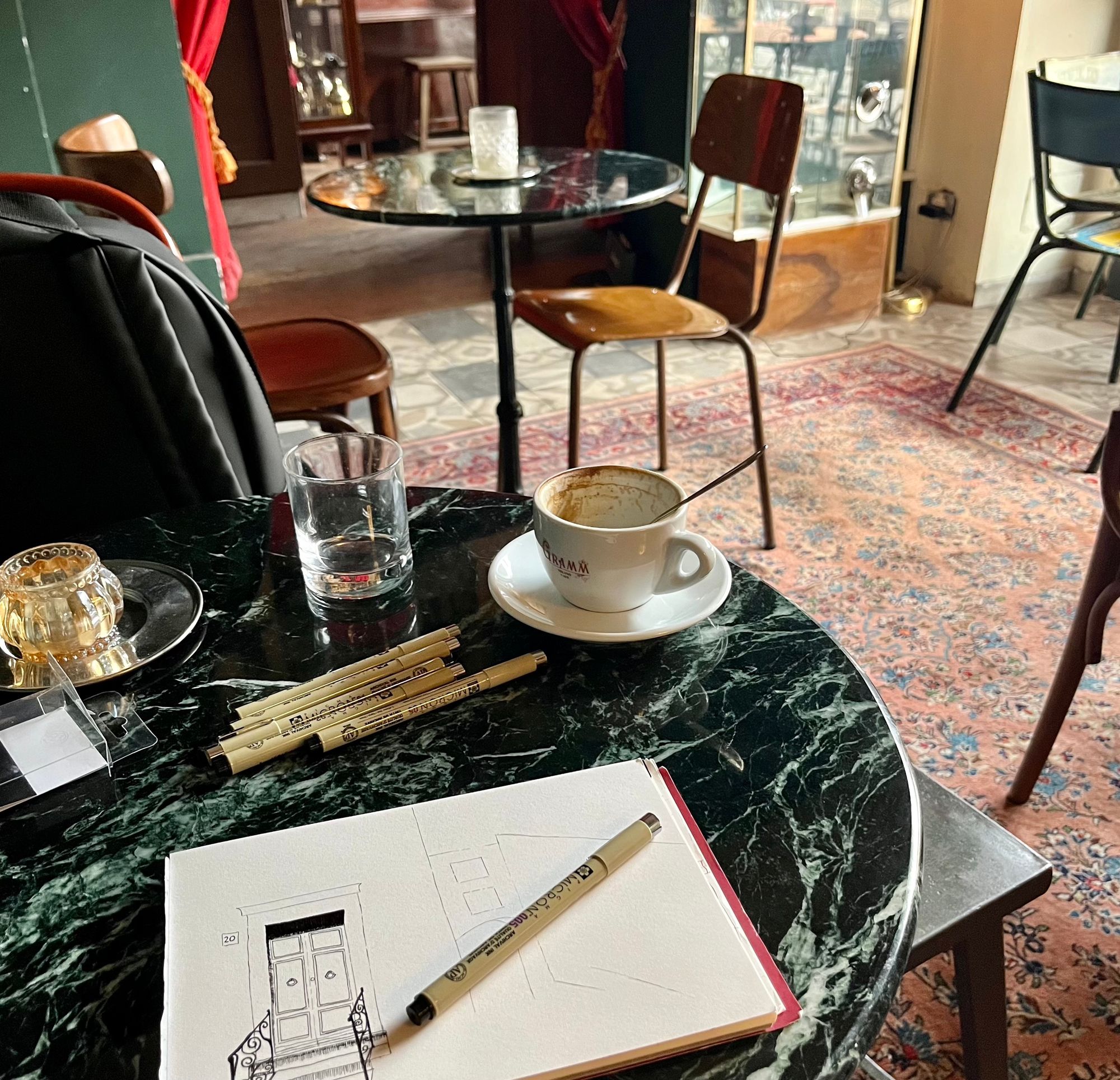 The interior of Gramm cafe in Navigli, Milan. A pen-sketched drawing lies on a marble table above a Persian rug. 