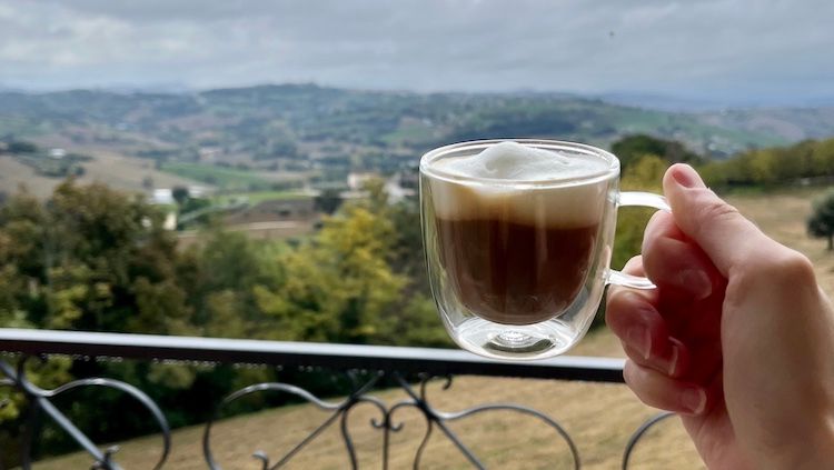 A clear glass holds a foamy espresso shot in the foreground of the Macerata foot hills.