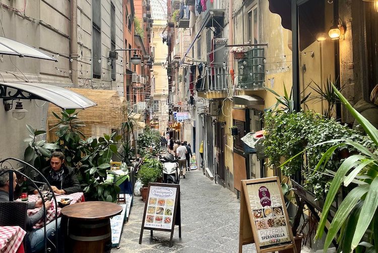 A busy street in Naples during the winter. Lights are strung above the shops and clothes hang on the balconies.