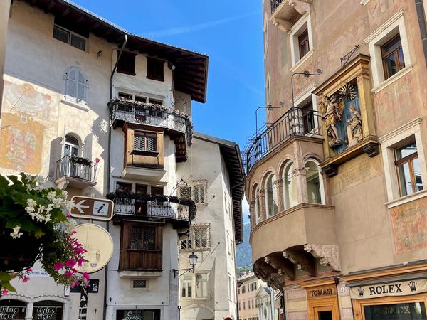 An Italian street with historic buildings, frescoes, balconies, and a Rolex shop under a clear blue sky.